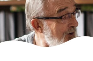 Idoso de óculos conversa com outras pessoas em ambiente de trabalho, com estantes e pastas ao fundo.