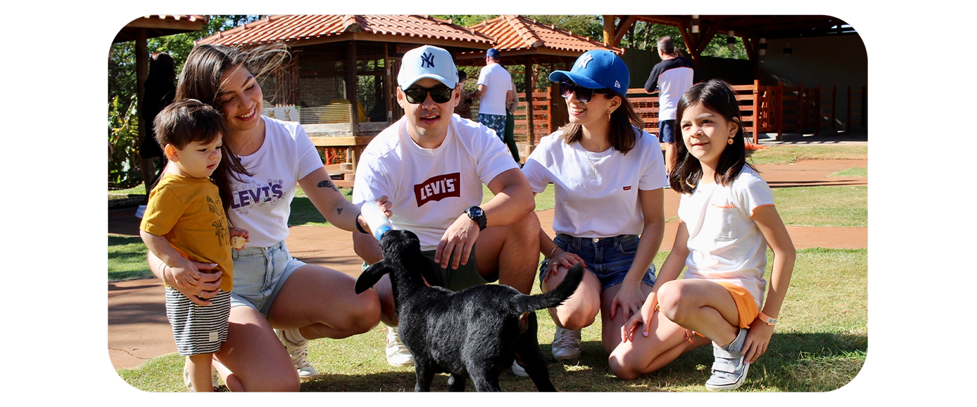 Família alimentando um filhote de cabra em área verde com estrutura rústica ao fundo.