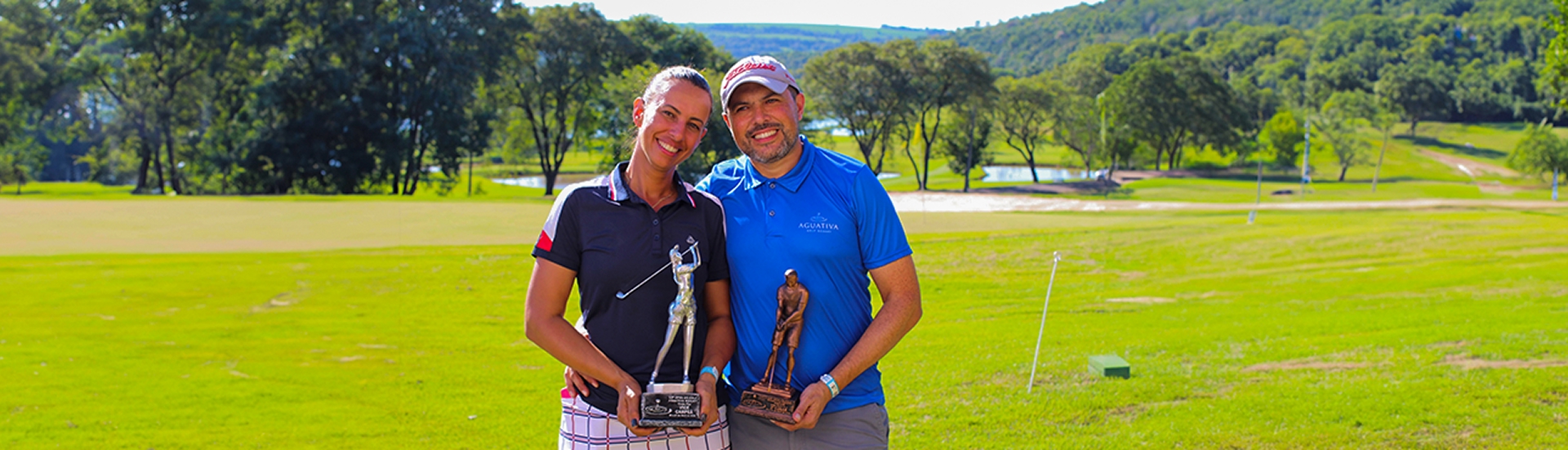 Casal de golfistas sorridentes posa com troféus em campo de golfe verde e arborizado do Aguativa Golf Resort.