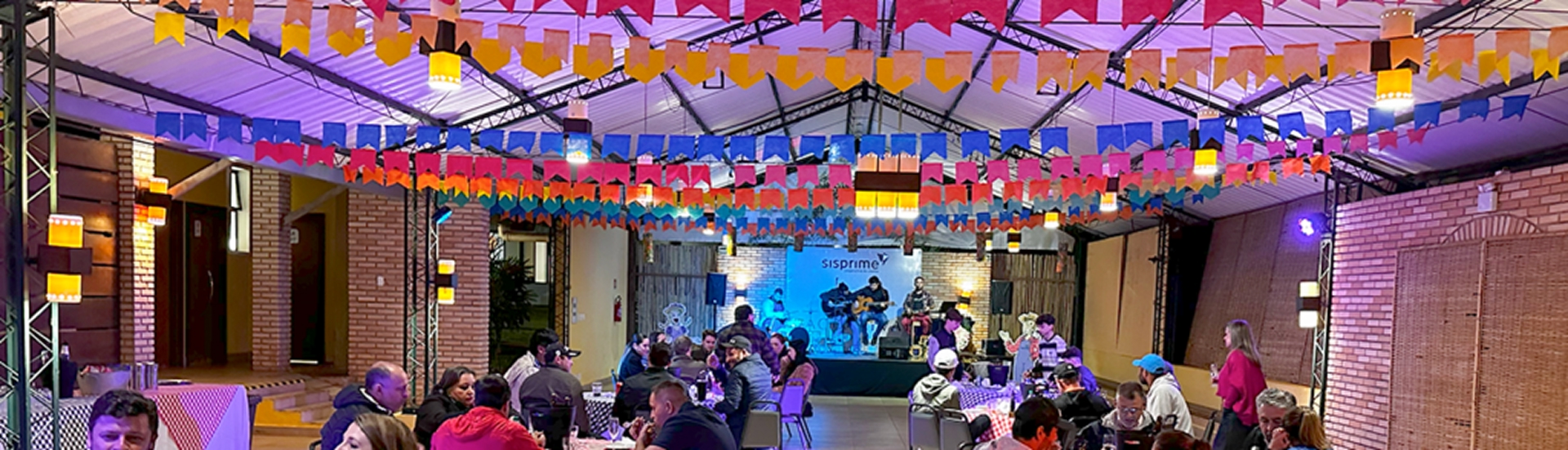 Festa junina indoor com decoração típica de bandeirinhas coloridas no teto, público sentado em mesas e apresentação musical ao vivo no palco no Aguativa Golf Resort.