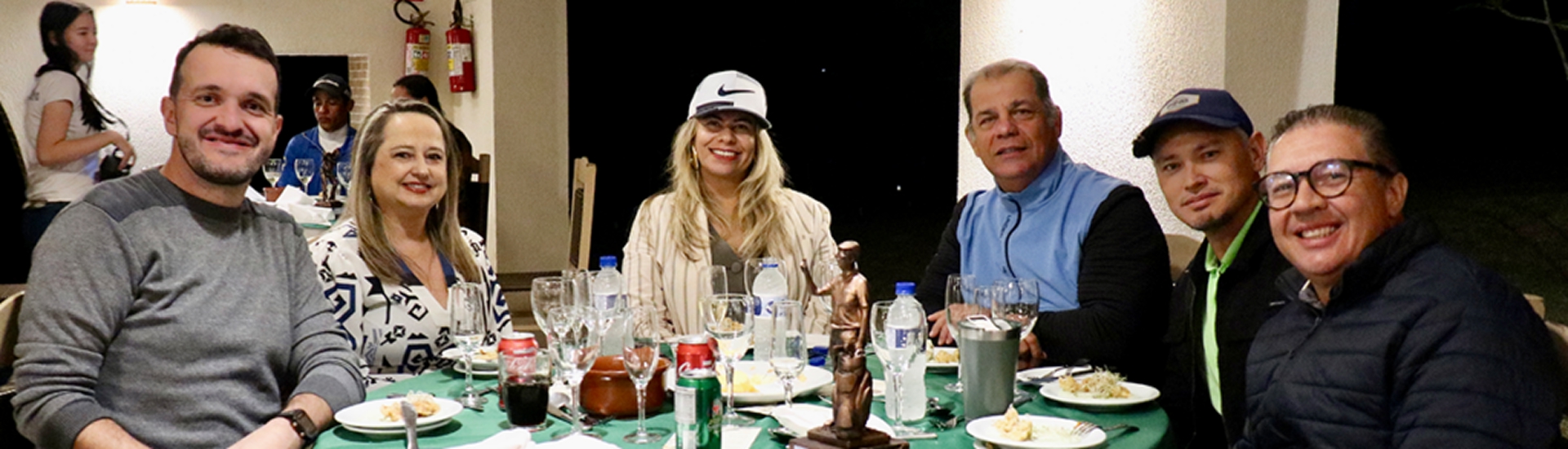 Grupo de sete pessoas sorrindo ao redor de uma mesa posta durante jantar no Aguativa Golf Resort, com troféus expostos e ambiente descontraído.