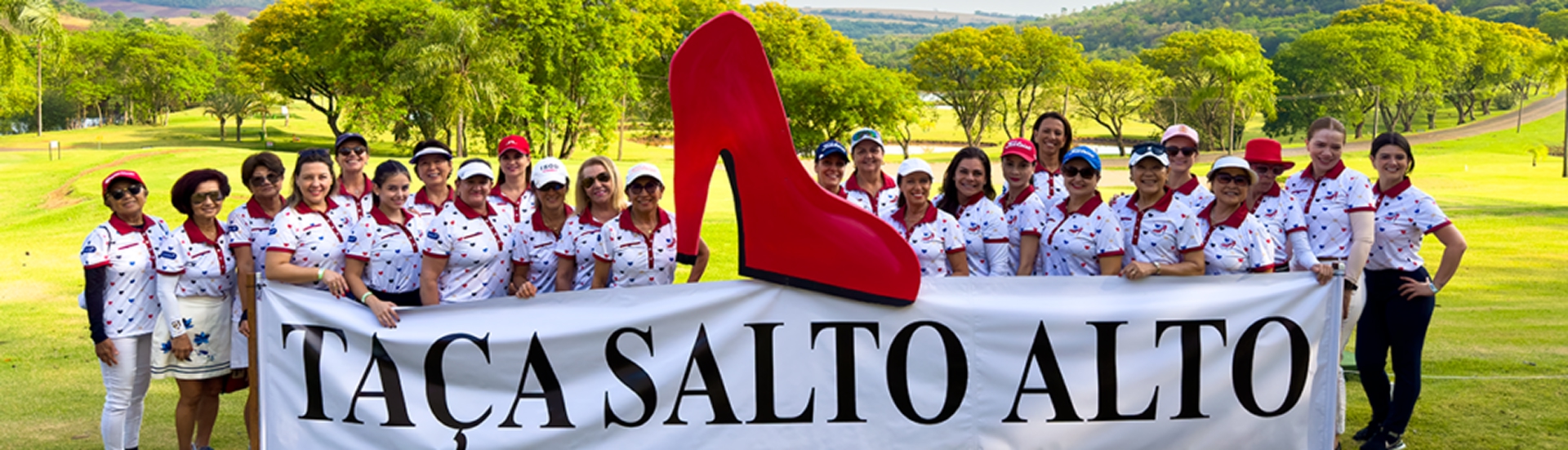 Grupo de mulheres golfistas sorrindo atrás de uma faixa com os dizeres "Taça Salto Alto", acompanhada de uma grande escultura de salto alto vermelho, em um campo de golfe arborizado.