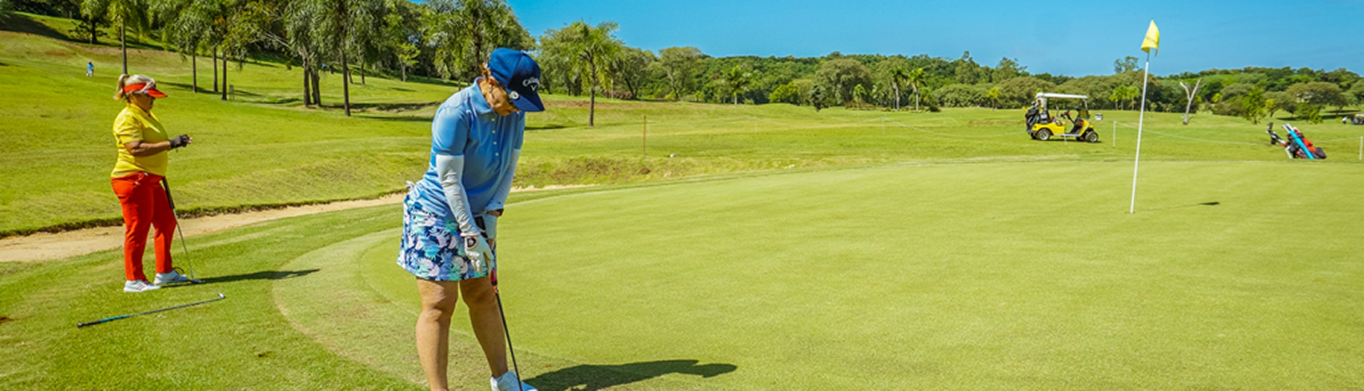 Duas mulheres jogando golfe em um campo gramado sob céu azul; uma se prepara para o putt enquanto a outra observa, com carrinho de golfe ao fundo.