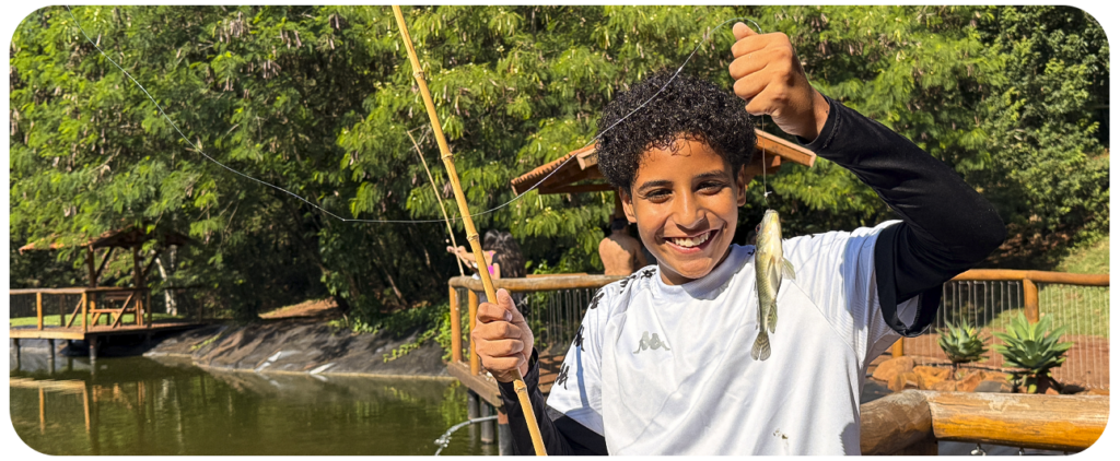 Garoto sorrindo enquanto segura pela linha um peixe que acabou de pescar.